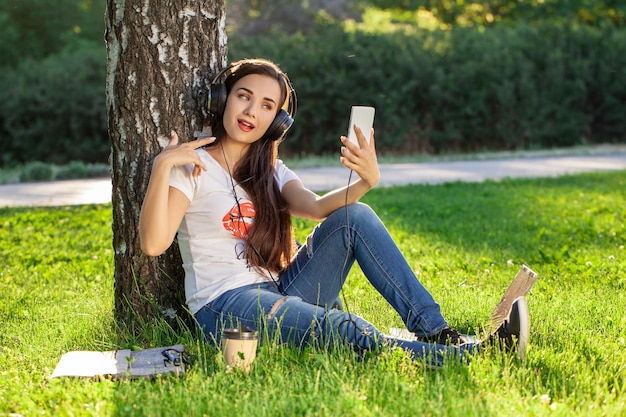 Mulher relaxa com fones de ouvido, ouvindo música, sentado na grama do parque. Mulher jovem com fones de ouvido fazendo selfie em seu smartphone