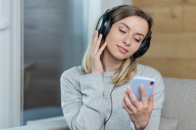 Mulher relaxa em casa e ouve música com grandes fones de ouvido usando o celular