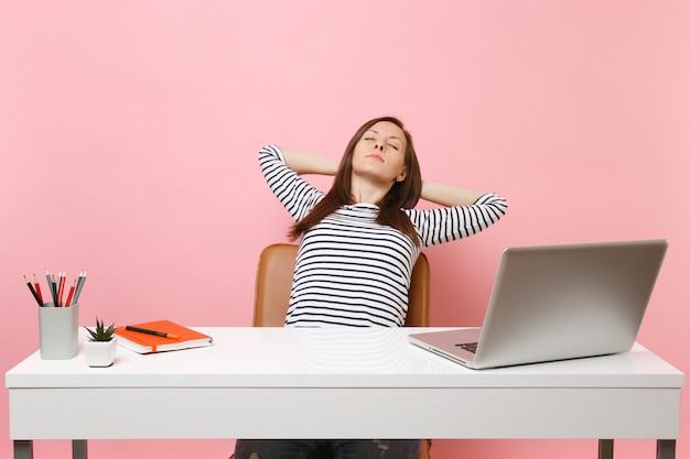 Mulher relaxada com os olhos fechados, segurando as mãos atrás da cabeça, sentada no trabalho em uma mesa branca com um laptop pc contemporâneo