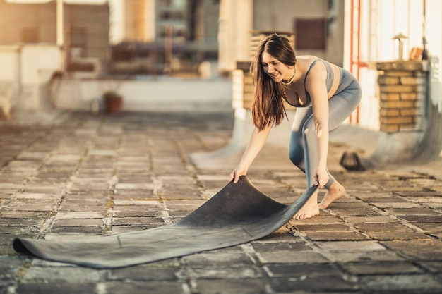 Mulher relaxada está preparando colchonete para praticar ioga em um terraço ao pôr do sol.