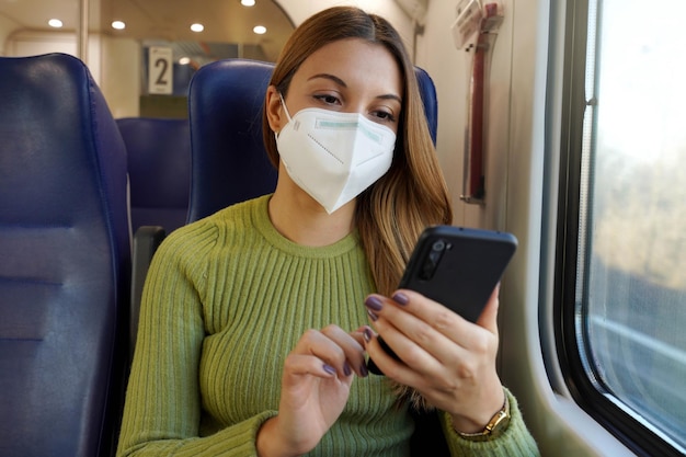 Mulher relaxada no trem usando máscara facial médica usando aplicativo de telefone inteligente. Viajante com máscara protetora viajando sentado em mensagens de texto de classe executiva no celular. Viaje com segurança no transporte público.