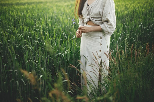 Mulher relaxando no campo de trigo na visão cortada à noite Elegante jovem fêmea em vestido rústico segurando flores silvestres nas mãos na zona rural de verão Momento atmosférico tranquilo