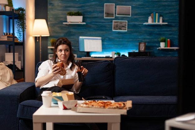 Mulher relaxando no sofá segurando uma garrafa de cerveja enquanto come um hambúrguer delicioso