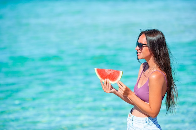 Mulher relaxante na praia num dia de verão, sorrindo com uma fatia de melancia na mão