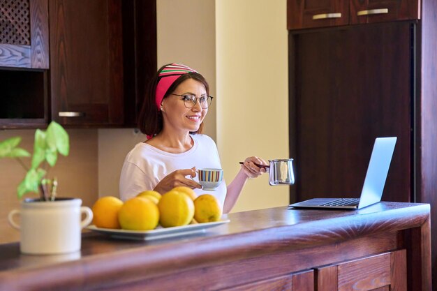 Mulher relaxante sorridente com café da manhã com cezve turk e laptop na cozinha em casa