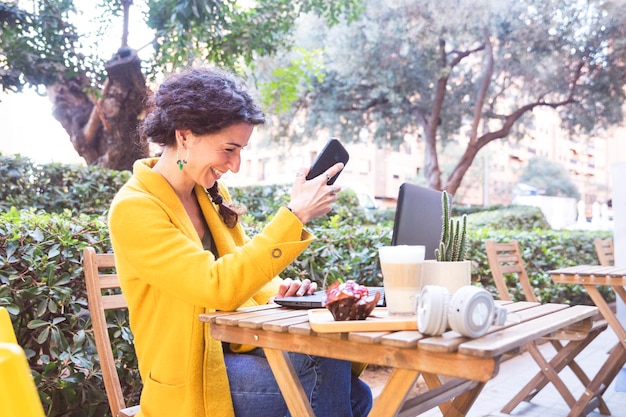 Foto mulher ri na frente de um computador com um celular na mão em um terraço