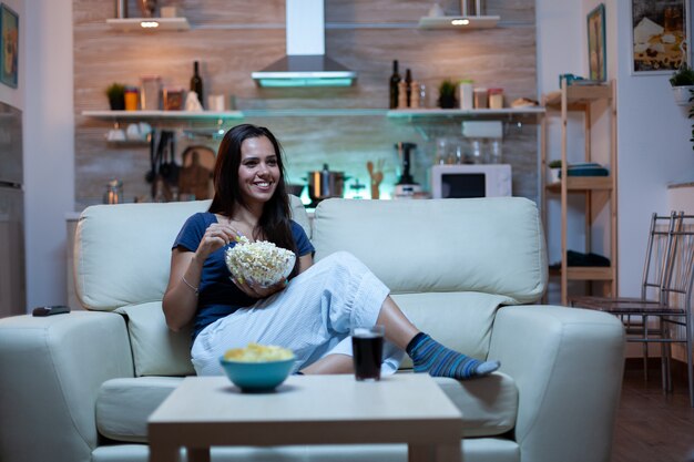 Mulher rindo assistindo tv e come lanches. jovem feliz, animada, divertida e sozinha em casa, curtindo a noite sentada em um sofá confortável, vestida de pijama, comendo pipoca em frente à televisão