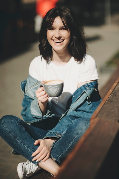 Mulher rindo com cabelo escuro em roupas jeans simples, sentada com as pernas cruzadas em um banco de madeira ao ar livre e segurando um copo com bebida na mão, olhando para a câmera