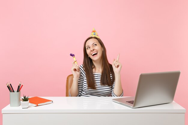 Mulher rindo com chapéu de festa de aniversário com cachimbo, comemorando enquanto se senta e trabalha na mesa branca com laptop pc isolado em fundo rosa pastel. Conceito de carreira empresarial de realização. Copie o espaço.