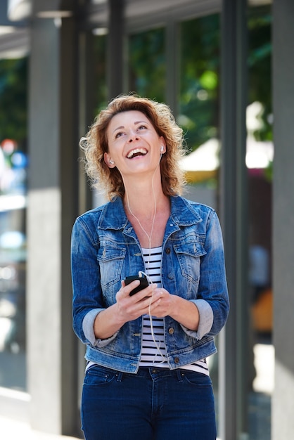 Mulher rindo com telefone e fones de ouvido andando na cidade