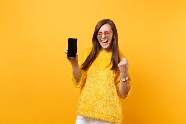 Mulher rindo em copos de coração cerrando o punho como vencedor segurando o telefone móvel com tela preta vazia em branco isolada no fundo amarelo brilhante. Estilo de vida de emoções sinceras de pessoas. Área de publicidade.