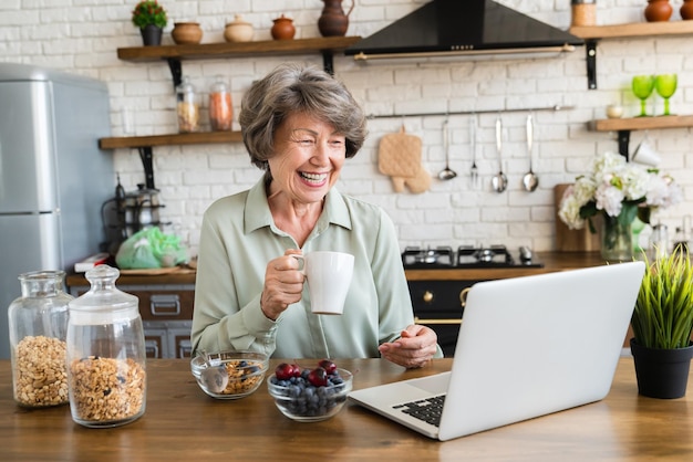 Mulher rindo vendo vídeo de filme em laptop na cozinha de casa enquanto faz café da manhã saudável