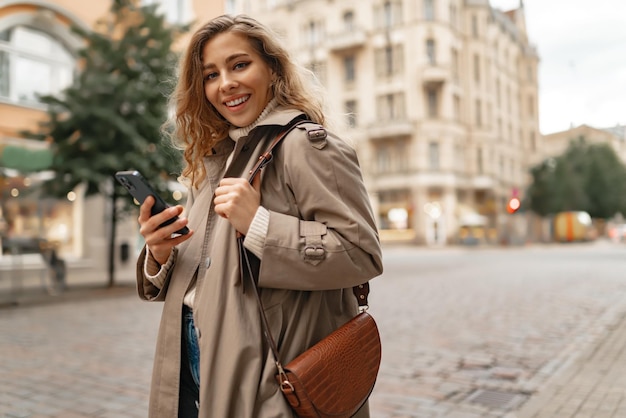 Mulher rizada sorridente vestindo um casaco quente caminhando pela rua e usando seu telefone
