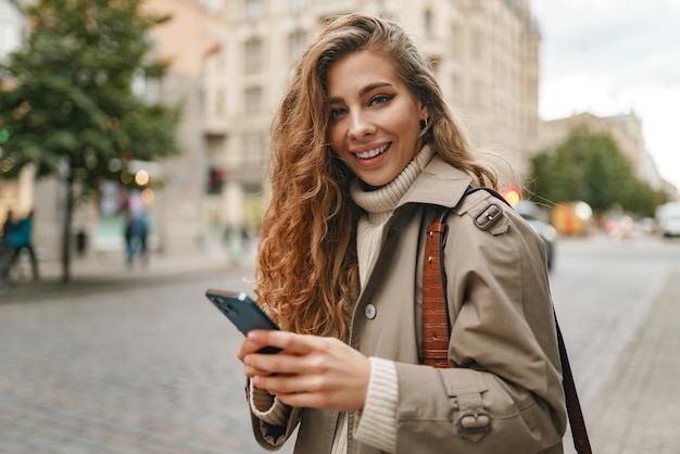 Mulher rizada sorridente vestindo um casaco quente caminhando pela rua e usando seu telefone