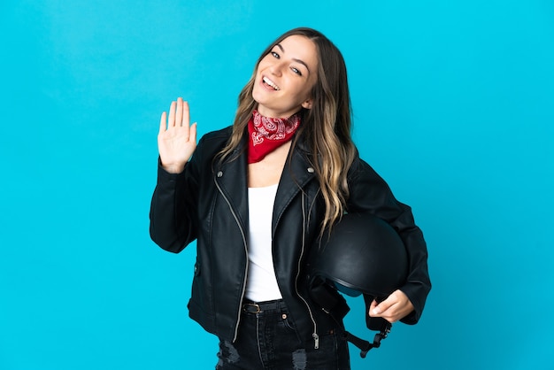 Mulher romena segurando um capacete de motociclista isolado na parede azul e saudando com a mão com expressão feliz