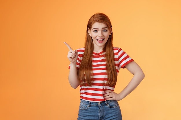 Mulher ruiva animada e simpática discute nova tendência apontando para o canto superior esquerdo sorrindo e conversando alegremente ...