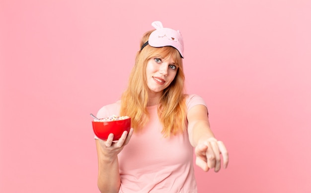 Mulher ruiva apontando para a câmera escolhendo você de pijama e segurando uma tigela de flocos