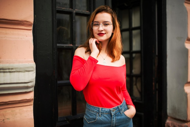 Mulher ruiva atraente em óculos usa blusa vermelha e saia jeans posando na rua contra a velha porta de madeira