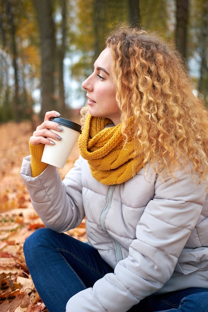 Mulher ruiva bonita e cacheada sentada nas folhas de outono, segurando café e apreciando a vista do parque de outono