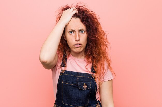 Mulher ruiva bonita jovem ruiva vestindo um jeans dungaree cansado e muito sonolento, mantendo a mão na cabeça.