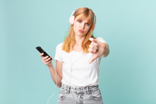 Mulher ruiva bonita se sentindo mal, mostrando os polegares para baixo e ouvindo música com fones de ouvido