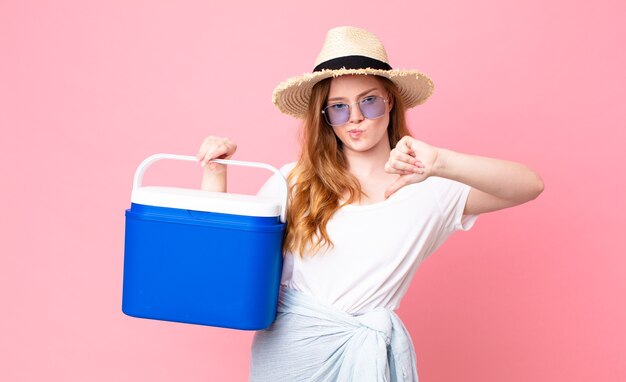 Mulher ruiva bonita se sentindo mal, mostrando os polegares para baixo e segurando uma geladeira portátil de piquenique