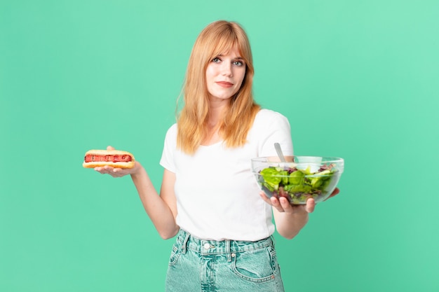 Mulher ruiva bonita segurando uma salada e um cachorro-quente. conceito de dieta