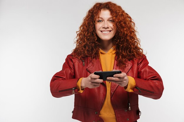 Foto mulher ruiva caucasiana de 20 anos vestindo uma jaqueta de couro, sorrindo e segurando o celular isolado na parede branca