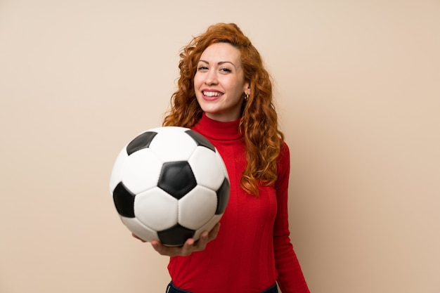 Mulher ruiva com camisola de gola alta, segurando uma bola de futebol