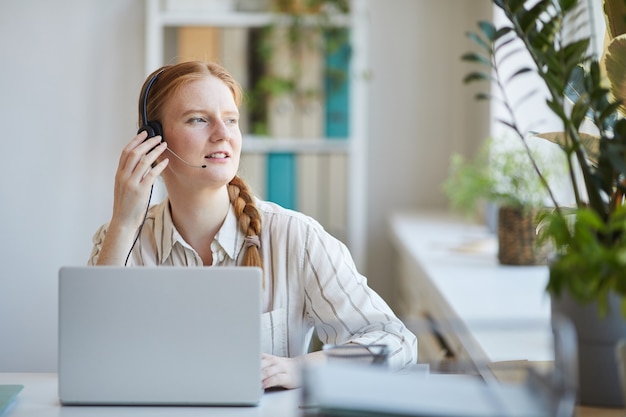 Mulher ruiva com fones de ouvido, sentada no local de trabalho, trabalhando em uma central de atendimento