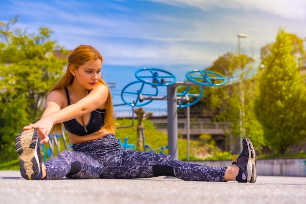 Mulher ruiva com legging roxa e macacão preto se exercitando nas máquinas, fazendo alongamento sentada no chão, esporte na cidade