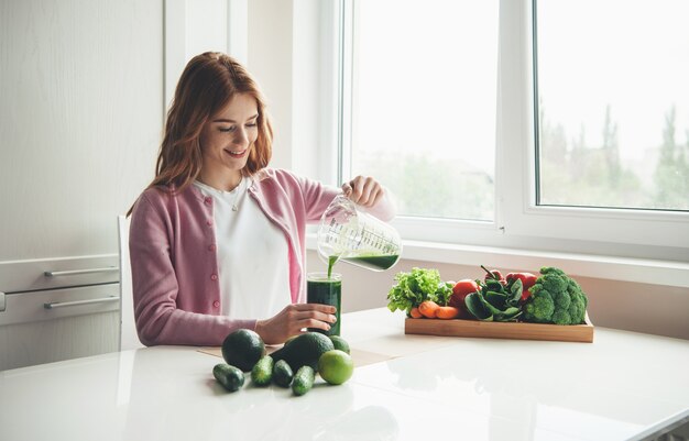 Mulher ruiva com sardas fazendo suco verde de vegetais frescos em casa colocando em um copo