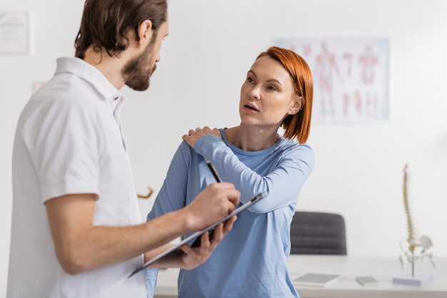 Mulher ruiva com um ombro ferido a falar com um fisioterapeuta a escrever um diagnóstico no quadro