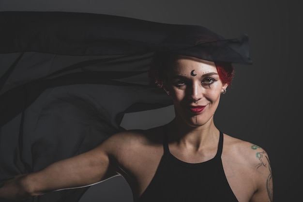 Mulher ruiva com um véu preto na cabeça em um estúdio escuro Uma garota com um brinco no nariz sorri maliciosamente Maquiagem de bruxa Traje de Halloween