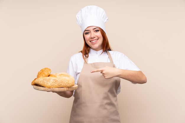 Mulher ruiva de adolescente em uniforme de chef. Padeiro feminino segurando uma mesa com vários pães e apontando