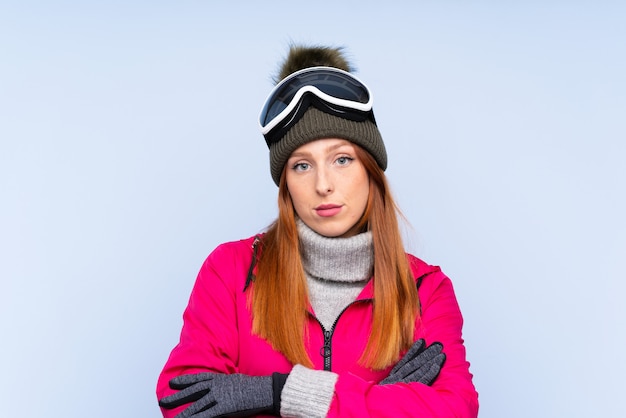 Mulher ruiva de esquiador com óculos de snowboard sobre parede azul isolada, mantendo os braços cruzados