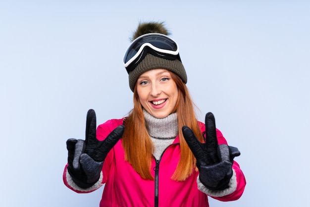Mulher ruiva de esquiador com óculos de snowboard, sorrindo e mostrando sinal de vitória