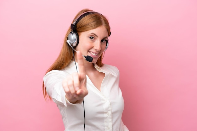 Mulher ruiva de telemarketing trabalhando com um fone de ouvido isolado no fundo rosa mostrando e levantando um dedo