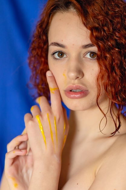 Mulher ruiva encaracolada com as mãos em tinta amarela Olha para a câmera sobre um fundo azul. Fotografia conceitual para arte ou blog feminino