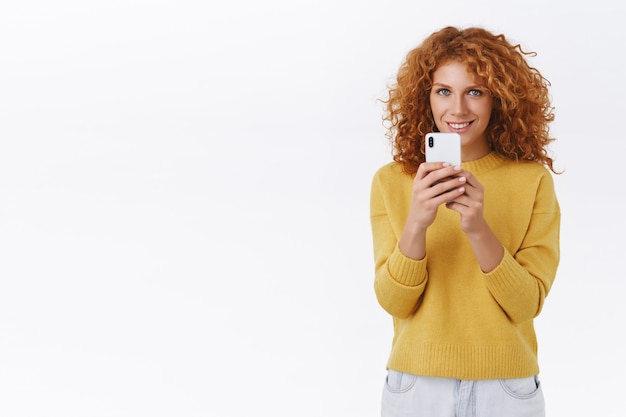 Mulher ruiva encaracolada divertida em um suéter amarelo, segurando smartphone e sorrindo satisfeita, gravar vídeo, tirar foto, atirar em um amigo para rede social ou assistir a um show incrível, parede branca