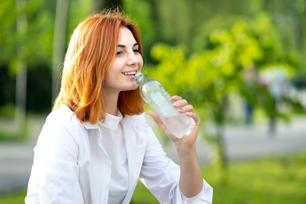 Mulher ruiva feliz e sedenta bebendo água mineral fresca no verão ao ar livre.