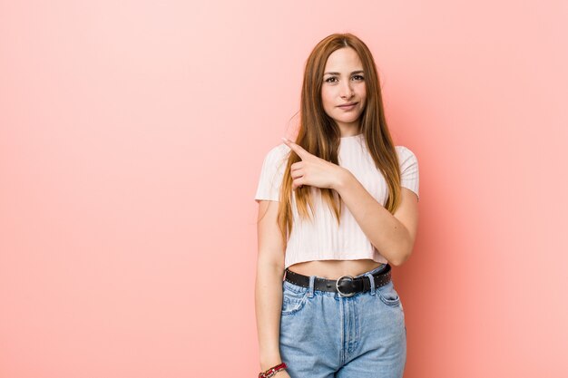 Mulher ruiva jovem gengibre contra uma parede rosa sorrindo e apontando de lado, mostrando algo no espaço em branco.