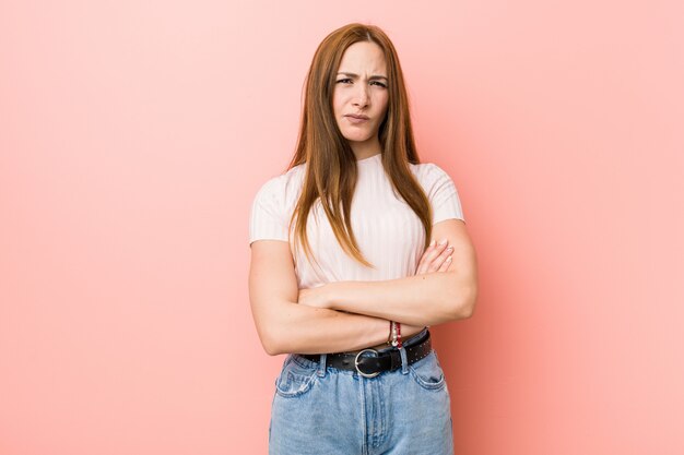 Mulher ruiva jovem ruiva contra um rosto carrancudo parede rosa em desagrado, mantém os braços cruzados.