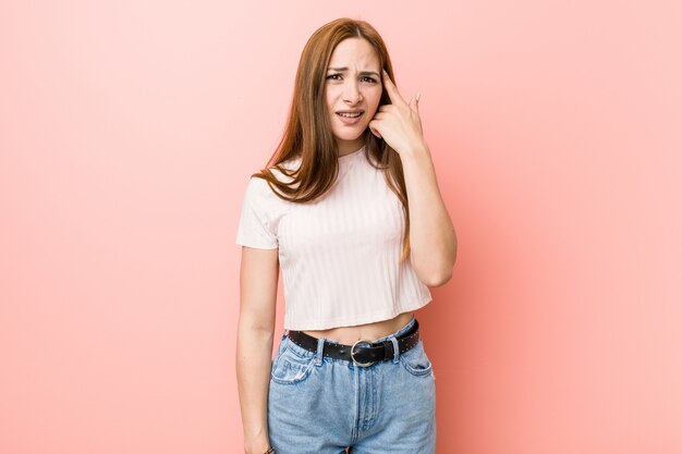 Mulher ruiva jovem ruiva contra uma parede rosa, mostrando um gesto de decepção com o dedo indicador.