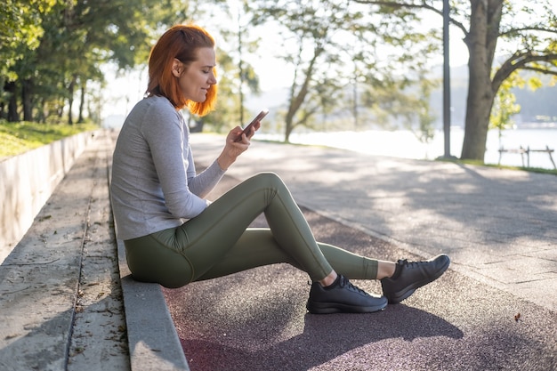 Mulher ruiva magra em roupas esportivas usando telefone celular no parque saúde, bem-estar, fitness, estilo de vida