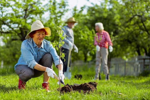 Mulher ruiva. Mulher aposentada ruiva bonita usando chapéu, cavando um terreno perto de uma árvore depois de plantá-la