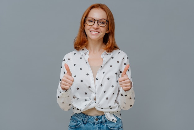 Mulher ruiva positiva com um sorriso gentil mostra os polegares, demonstra como ou gesto de aprovação