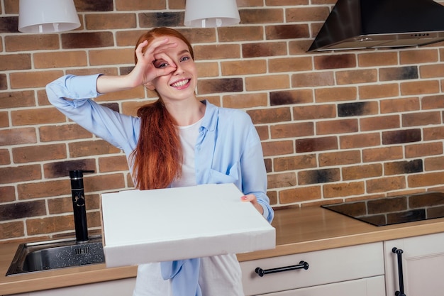 Mulher ruiva segurando caixa com pizza na cozinha em casa