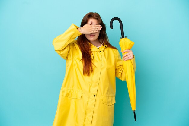 Mulher ruiva segurando um guarda-chuva isolado em um fundo azul, cobrindo os olhos com as mãos. Não quero ver nada