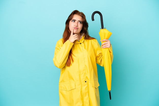 Mulher ruiva segurando um guarda-chuva isolado em um fundo azul, tendo dúvidas enquanto olha para cima